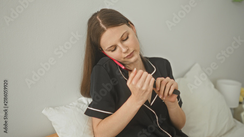 Fotografía Woman filing nails with an emery board on a bed while holding a pink smartphone to her ear in a bedroom; relaxation self care