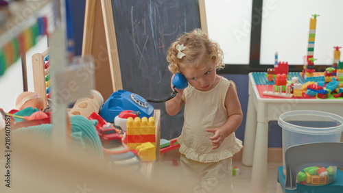 Little blonde girl holds blue toy telephone to her ear and dials beside a chalkboard and table of blocks in a playroom building; curiosity and play.