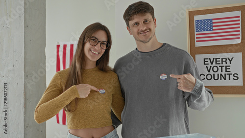 Young woman points finger to voted sticker on chest beside partner at ballot box in polling station building; civic engagement pride.