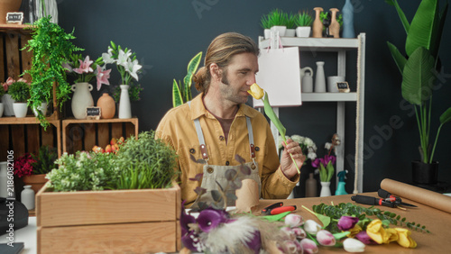 Фотография Man florist with apron sniffs a single yellow tulip beside a workbench strewn with flowers, tools and wrapping paper in a studio; serenity