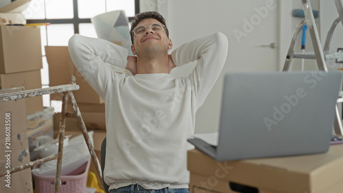 Konstfotografi Man at a makeshift desk with laptop and cardboard boxes reading papers then leaning back with hands behind head in a building; relief settling