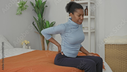 Obraz na plátně Woman sitting on bed with hand on lower back, grimacing with backache while shifting weight in building bedroom near plant and wicker basket; pain recovery