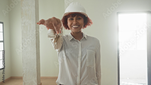 Картината върху платно Woman holding keys with outstretched hand in an empty building room, smiling while wearing hardhat and striped shirt as architect presenting access; homeowner pride