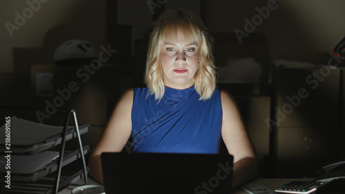 Obraz na plátně Woman in blue top typing on laptop while holding a mug and closing her eyes, surrounded by parcels and boxes in a building; late night fatigue