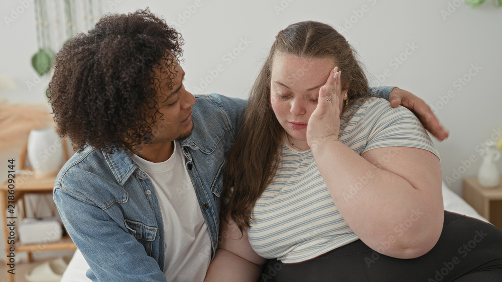 Obraz premium Woman holds hand to forehead on bed while man wraps an arm around her shoulder in a bedroom, leaning close in a consoling embrace; comfort support intimacy.