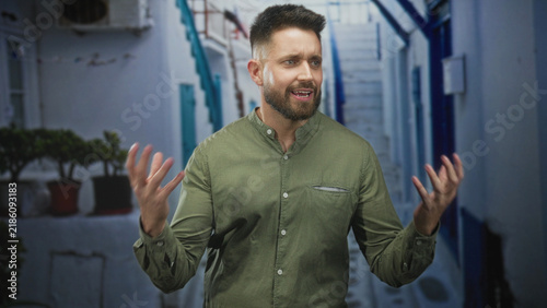 фотографія Man with beard gesturing with hands and closed eyes on a narrow sunlit street with white buildings and potted plants; frustration plea