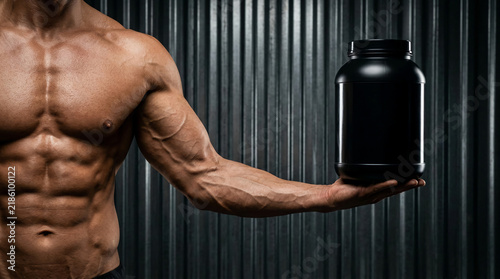 Muscular shirtless bodybuilder holding a blank black protein powder tub mockup for sports nutrition against a dark corrugated metal background fitness supplement advertising and extreme gym strength