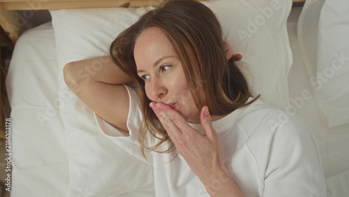 Obraz na płótnie Woman on bed covering mouth with hand, smiling and gazing sideways, bare hand visible, white sheets and wooden headboard in a bedroom inside a building; thoughtful intimacy