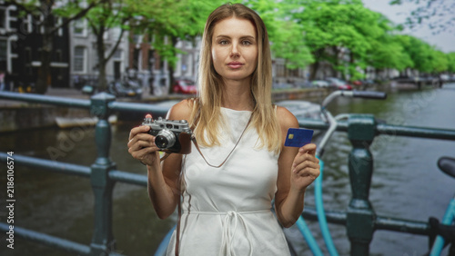 Obraz na plátně Woman holds vintage camera and creditcard on canal street bridge, facing forward with neutral gaze; confidence