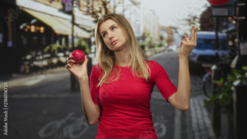 Photography Blonde woman in a red top holding a pomegranate in her right hand while shrugging and rolling eyes on a street lined with cafes and a van; indifference doubt