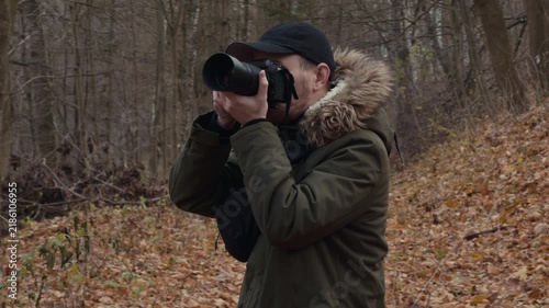 Photographer holding long telephoto lens to eye and framing view in autumn woodland, tight eye-level close-up showing hand grip, viewfinder focus, and warm natural light