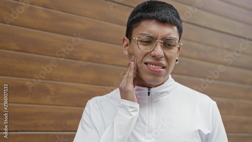 Принти на полотні Man hand on cheek wincing for toothache beside building wall, wearing white zippered top and glasses; toothache pain recovery