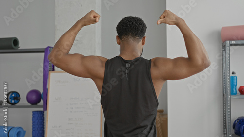Fotografie Man flexes both biceps, facing away in a training studio with workout gear and a visible whiteboard; strength confidence