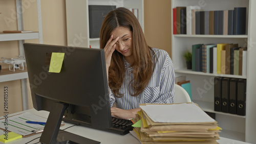 Fotografie Woman typing at desktop computer with piles of files and paperwork, touching temple and wearing glasses at office; stress overload