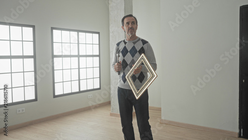 Man holds hammer and diamond picture frame in hand while looking up at a wall in an empty building; contemplation home repair.