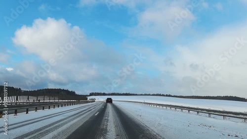 A car moves along a snowy road in a remote area. Snow covers the road and fields, creating a winter landscape. The sky is mostly clear with some clouds. There are wind turbines and a visible sign 