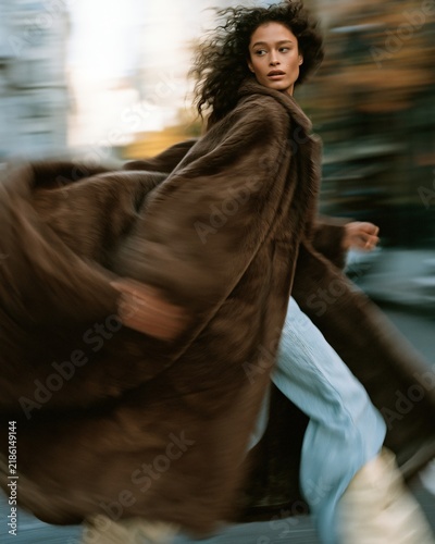 A model walks quickly on a city street wearing a brown fur coat and jeans