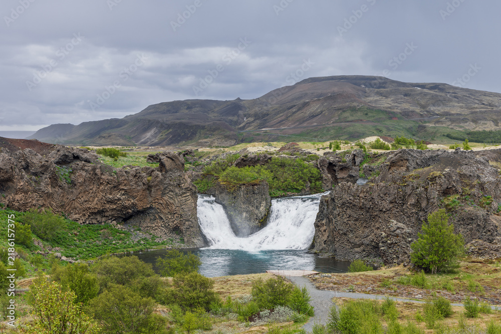 Fototapeta premium Hjalparfoss waterfall in the volcanic landscape of Thjorsardalur in wild Icelandic scenery. Gentle daylight preserves subtle earth and green tones. The shrubs define this vivid volcanic landmark.
