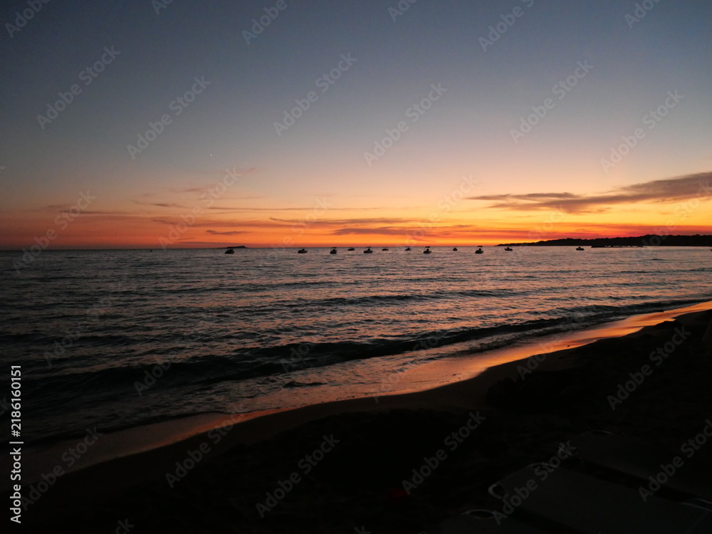 Obraz premium Coucher de soleil paisible sur l'océan avec des teintes orange et roses se reflétant sur l'eau et de petits bateaux en silhouette à l'horizon.