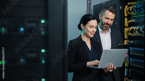 Smiling female IT specialist working on laptop while standing next to male colleague, professional engineers monitoring mainframe system in cloud data center server room