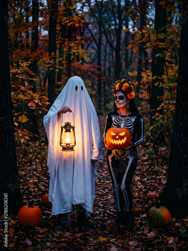 Ghost and skeleton girl with glowing jack-o-lanterns in a dark autumn forest at night
