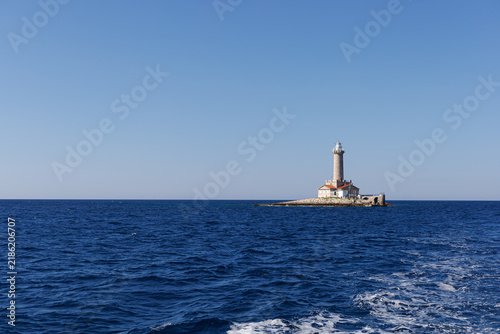 Fotografía Distant lighthouse marks a small rocky islet beyond the wake of a moving boat with broad copy space