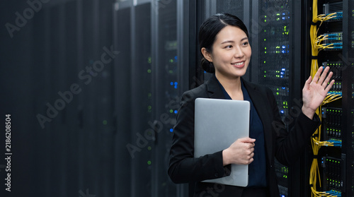 Smiling Asian female IT engineer holding laptop and waving hand in corporate cloud data center, professional system administrator standing near mainframe server rack equipment