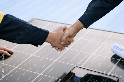 Two businesspeople shake hands over a solar panel table with a smartphone, partnership, renewable energy investment and clean technology agreement.
