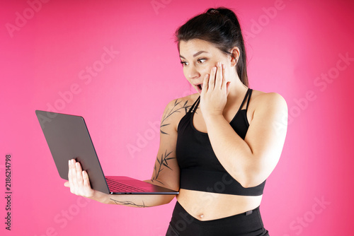 A surprised athletic young woman looks at the screen of her laptop and is amazed  studio with pink background