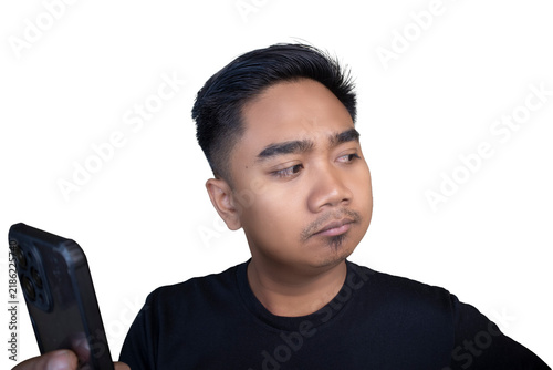 Fotografie Skeptical Indonesian man wearing a black t-shirt looking away from his mobile phone with a pouting and doubtful expression against a white background