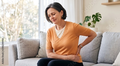 A middle-aged woman is sitting on a couch and holding her lower back in pain.