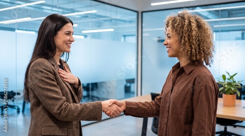 Two business women shaking hands in a modern office, smiling and looking at each other
