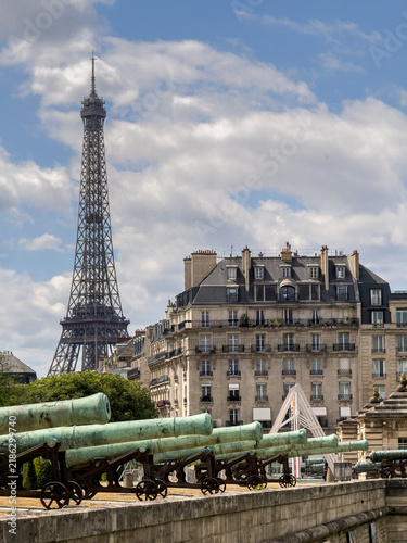 Rows of weathered bronze cannons stand guard at Les Invalides, with the Eiffel Tower rising above Parisian rooftops in the distance.