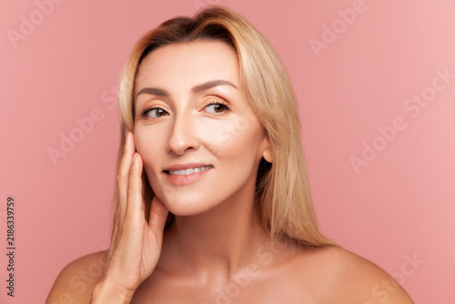 Radiant woman looking away and touching cheek with fingers for facial lifting and hydration concept isolated on pink background