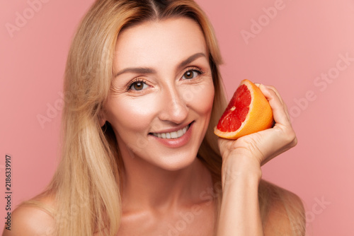 Radiant woman squeezing slice of red grapefruit near face, demonstrating refreshing citrus therapy and organic facial treatment isolated on pink background