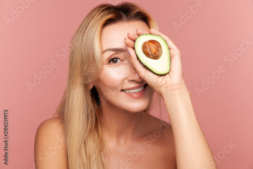 Playful woman covering eye with half of green avocado, showing organic ingredient for homemade face masks and nourishment isolated on pink background