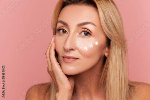 Mid-adult woman with blonde hair gently touching her face while posing with three moisturiser dots under eye isolated on pink background