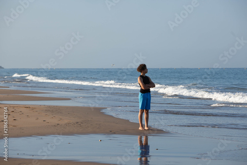 ​Young Man Standing Reflectively on Beach Looking Out at Ocean, Low Angle Shot. A barefoot young man with curly dark hair stands alone on a sandy beach.
