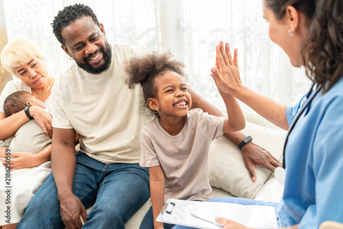 Close up of a young girl laughing as she high fives the home health nurse, her father laughing beside her on the sofa.