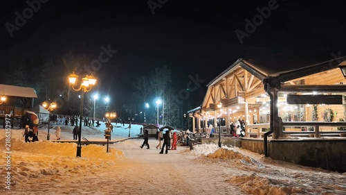 People are gathered at a ski resort during the night. Snow covers the ground, and warm lights illuminate the area. Some individuals are skiing or snowboarding, while others chat and walk near the lodg