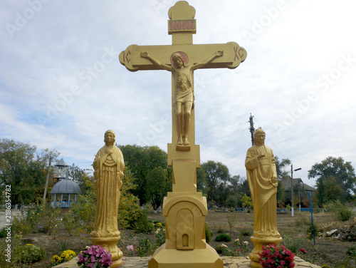 Tiraspol, Moldova - October 15, 2021: An Orthodox cross with the crucifixion of Jesus Christ, mounted on a pedestal, flanked by sculptures of saints, and surrounded by a well-kept garden of flowers