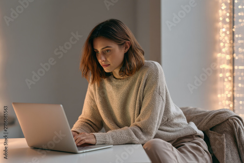 Young woman working on a laptop at home in cozy sweater  
