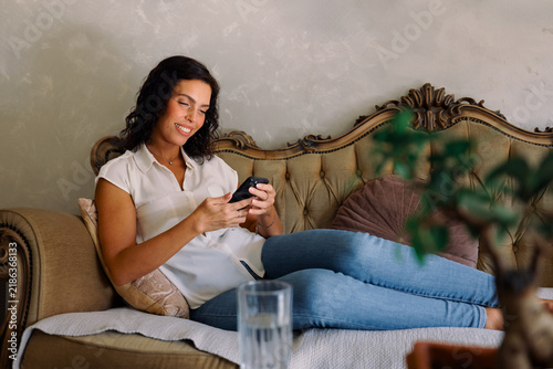 Fotografía Happy woman lounging on a vintage sofa, smiling as she scrolls and texts on her