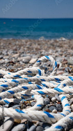 A close-up of white rope with blue accents laid on a pebbly beach, with the tranquil sea in the background under a clear blue sky.