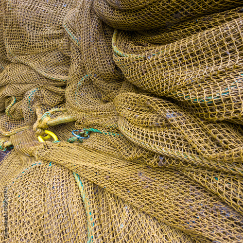 A detailed close-up of piled, textured yellowish-brown commercial fishing nets on a deck, featuring complex mesh patterns, knots, and a visible yellow hardware ring.