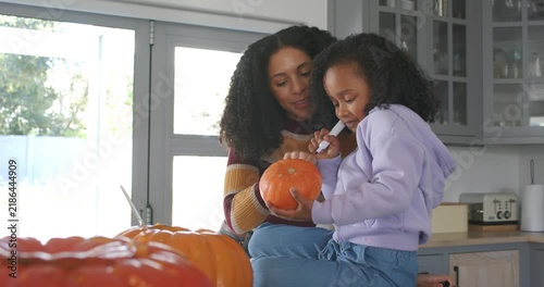 African American mother handing pumpkin, guiding child tracing with pen on kitchen counter for fall