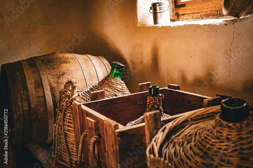 Bild auf Leinwand Close-up of wicker-covered demijohn bottles and a wooden barrel in a rustic cellar with warm sunlight