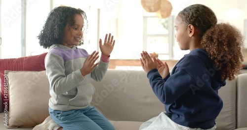 African American girls kneeling on couch raising hands playing clap game high-fiving near window