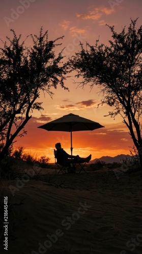 Silhouette of a person sitting in a camping chair under a parasol, enjoying a scenic desert sunset.