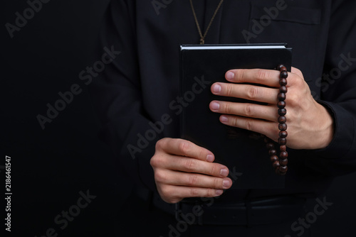 Fotografie Priest in cassock with rosary and Holy Bible on black background, closeup